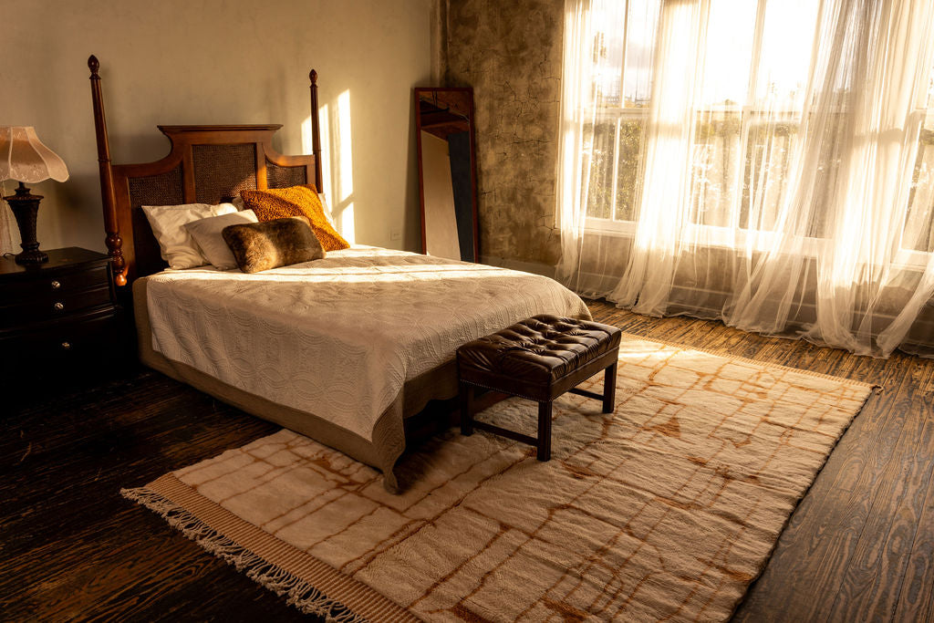 Light is peering into a beautiful room with a soft, plush handwoven Moroccan rug.  The rug is situated in a stunningly placed interior underneath a bed and leather ottoman.  The wind is blowing through the window and the curtains accentuate the naturally woven rug. 